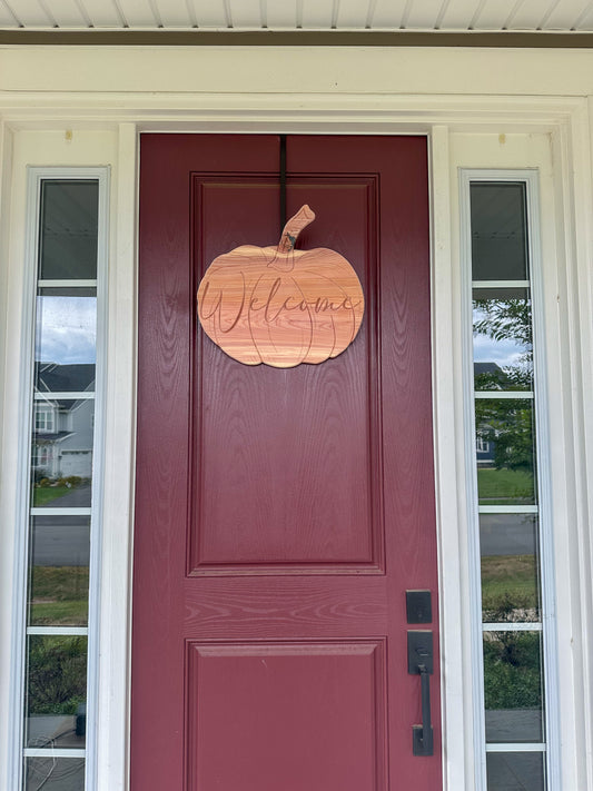 Wooden pumpkin-shaped 'Welcome' sign on a maroon door with glass panels.